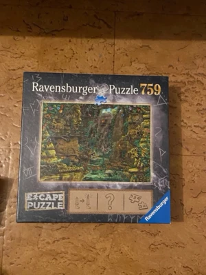 Ravensburger Escape Puzzle 759 bitar - Escape Puzzle från Ravensburger med 759 bitar. Motivet visar en mystisk djungel med vattenfall och gömda symboler. Kartongen är svart med gåtfulla tecken och blå detaljer. Perfekt för dig som gillar kluriga pussel och äventyr.