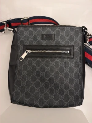 Gucci Messenger bag - Snygg svart axelväska från Gucci med klassiskt GG-mönster och detaljer i läder. Väskan har en framficka med dragkedja och en bred axelrem i rött och blått. Perfekt för dig som vill ha en stilren och lyxig accessoar.