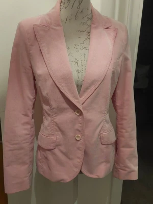 Ljusrosa kavaj med två knappar - Snygg ljusrosa kavaj med två knappar framtill och klassiska slag. Kavajen har två fickor med lock och är figursydd för en modern siluett. Tillverkad i ett mjukt tyg som ger en bekväm känsla. Lite liten i storleken.. Köpt på MQ.