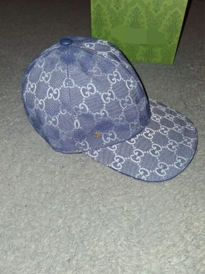 Blå Gucci keps med monogram - Snygg blå keps från Gucci med klassiskt GG-monogram över hela kepsen. Kepsen har böjd skärm och är tillverkad i denimliknande material. Perfekt för dig som vill sticka ut med en exklusiv accessoar. Lådan får man inte vid köpet.