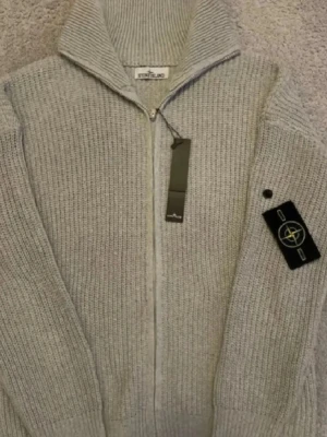 Grå stickad ziptröja från Stone Island - Snygg grå stickad ziptröja från Stone Island med hög krage och ribbstickad struktur. Tröjan har dragkedja framtill och klassisk logotyp-patch på ärmen. Perfekt för lager-på-lager och stilren look. Har både i färgen svart och grå