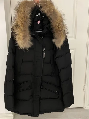 Svart dunjacka med äkta päls från Holliester - Snygg svart dunjacka från Holliester med stor huva och fluffig äktapäls i bruna toner. Jackan har dragkedja, knappar, flera fickor och quiltad design. Perfekt för kalla vinterdagar och riktigt varm.
