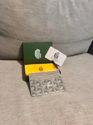 Grå kortfodral från G - Snyggt och stilrent kortfodral från G i grått med det klassiska G -mönstret i vitt. Tillverkat i canvas och läder, perfekt för kort och ID. Kommer med original ask och dustbag. En riktig statement-accessoar för dig som gillar exklusiva detaljer.