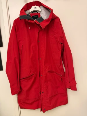 Röd parkasjacka från Didriksons - Röd parkasjacka från Didriksons med huva, dragkedja och tryckknappar framtill. Jackan har två stora fickor med dragkedja och justerbara ärmslut. Perfekt för kyliga dagar och har en sportig, stilren look.