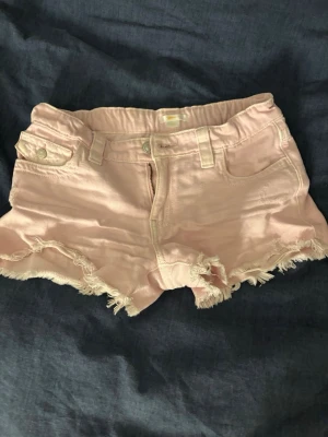 Ljusrosa fransiga jeansshorts - Säljer ett par ljusrosa jeansshorts med fransig kant och klassisk femficksdesign. Shortsen har bälteshällor, knappgylf och en bakficka med lock och knapp. Perfekta för varma dagar och en avslappnad stil.