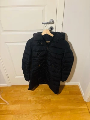 Svart lång dunjacka med huva - Snygg svart dunjacka med huva och dragkedja framtill. Storlek M. Jackan har en längre modell, två fickor med dragkedja och quiltad design. Perfekt för kalla vinterdagar och ger en stilren look. Passar dig som vill ha både värme och enkel stil. Äkta soklart, om är intresserande hojta till 😄