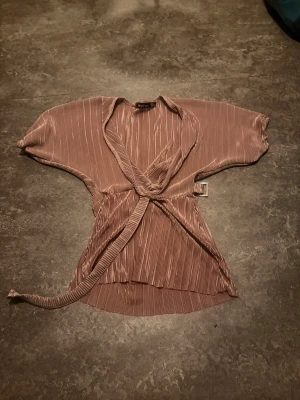 Brons omlottopp från Boohoo - Säljer en snygg brons omlottopp från Boohoo i glansigt, plisserat material. Toppen har korta vida ärmar och knyts i midjan för en trendig look. Perfekt att styla med jeans eller kjol för en cool outfit.