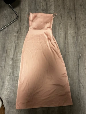 Lång ljusrosa satinklänning från vila - långklänning i ljusrosa satin med smala axelband och rak halsringning. Klänningen är lite öppen i ryggen och har även tyvärr fått en fläck på sig men syns inte när den är på osv.