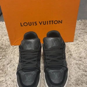 Louis Vuitton svarta sneakers - Svarta Louis Vuitton sneakers med klassiskt monogrammönster i mörkgrått. Skorna har rund tå, snörning och vit platt sula. Tillverkade i skinn med lyxig känsla och diskreta LV-detaljer över hela ovandelen.