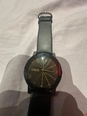 Svart klocka från GREK med guldiga detaljer - Stilren svart klocka från GREK med rund urtavla och guldiga visare samt detaljer. Klockan har ett svart band i syntetmaterial och minimalistisk design. Perfekt accessoar för att lyfta din outfit. Funkar ej!