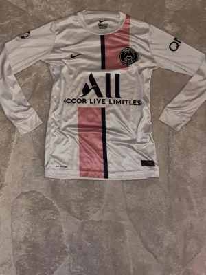 PSG långärmad matchtröja Nike - Snygg långärmad PSG matchtröja från Nike i vitt med rosa och svart rand på framsidan. Tröjan har klubbmärke, sponsortryck och rund hals. Perfekt för fotbollsträning eller att bära som supporter. Materialet är lätt och andas bra.