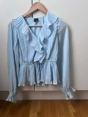 Ljusblå plisserad blus  - Supersöt ljusblå blus från vero moda  med plisserat tyg och volangdetaljer längs v-ringningen. Blusen har resår i midjan och vid ärmsluten för en snygg passform. Långärmad och tillverkad i ett lätt, luftigt material som ger en romantisk vibe.