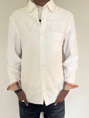 Vit skjorta med rutiga detaljer - Vit skjorta med klassisk krage och knäppning framtill. Manschetterna har snygga rutiga detaljer i beige, rött och svart. Skjortan har en bröstficka och är långärmad. Perfekt för en stilren look.