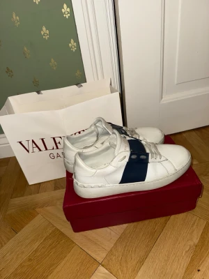 Valentino vita sneakers med blå detalj - Snygga vita sneakers från Valentino med bred mörkblå rem över sidan och coola nitar längs sulan. Skorna är i läder och har en klassisk låg modell som passar perfekt till streetwear eller en clean look. Obs färgen har lossnat ytte pytte lite.