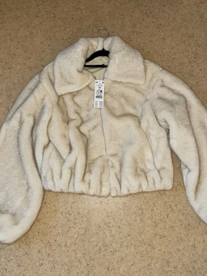 Beige kort pälsjacka med krage - Säljer en fluffig beige pälsjacka i kort modell med bred krage och dragkedja framtill. Jackan har ballongärmar och resår i midjan för en snygg siluett. Perfekt för dig som vill ha en trendig och varm vinterlook.