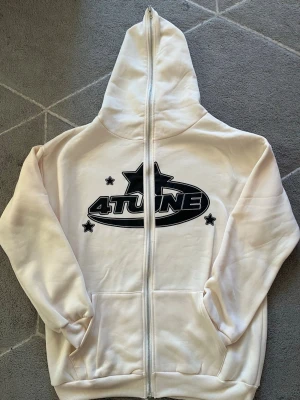 Beige hoodie med 4TUNE-tryck - Säljer en beige hoodie från 4TUNE med dragkedja och stor huva. Framsidan har ett svart 4TUNE-tryck med stjärnor och baksidan har spiralformat 4TUNE-motiv. Hoodien har kängurufickor och är gjord i mjukt material, perfekt för en avslappnad streetwear-look.