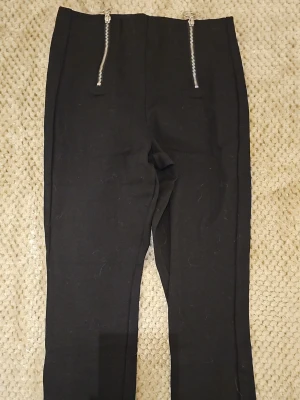 Svarta leggings med dragkedjor Lindex - Säljer ett par svarta leggings från Lindex i storlek S. De har två snygga silverfärgade dragkedjor framtill som detalj. Materialet är stretchigt och bekvämt, perfekt för en trendig och avslappnad look.