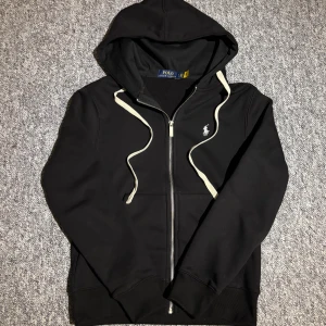 Svart hoodie från Polo Ralph Lauren S - Snygg svart hoodie med dragkedja från Polo Ralph Lauren. Klassisk design med vit broderad logga på bröstet och vita snören i huvan. Tillverkad i mjuk bomull, perfekt för en chill och stilren look. Pris kan diskuteras 😊