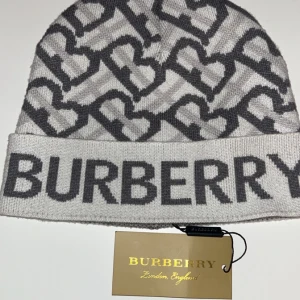 Gråvit Burberry mössa  - Snygg gråvit mössa från Burberry med tydlig logga framtill och mönster av sammankopplade bokstäver. Mjuk och stretchig stickad kvalitet, perfekt för att hålla värmen och ge en trendig look.