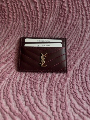 Vinröd YSL kortfodral i läder - Snyggt vinrött kortfodral från YSL i äkta läder med guldigt logomärke framtill. Fodralet har flera kortfack och dekorativa sömmar i matchande färg. Perfekt för att hålla koll på dina kort med stil.