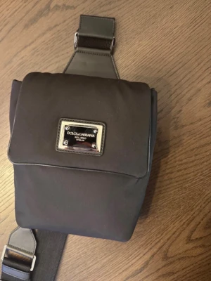 Svart axelväska Dolce & Gabbana - Snygg svart axelväska från Dolce & Gabbana med justerbar bred axelrem och metallplatta med logga på framsidan. Väskan är tillverkad i slitstark nylon och har en stilren, fyrkantig form som passar perfekt till en trendig look.