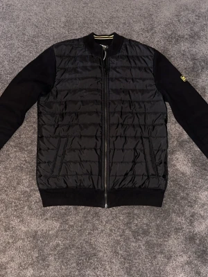 Svart cardiganjacka från Barbour - Snygg svart cardiganjacka från Barbour med quiltad front och stickade ärmar. Dragkedja framtill, ribbade muddar och en gul Barbour International-patch på ena ärmen. Perfekt för lager på lager och streetwear-vibe.