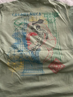 Casablanca grön t-shirt med print - Cool grön t-shirt från Casablanca med färgglatt grafiskt tryck på både fram- och baksidan. Motivet har en konstnärlig touch med ansikte, tennisspelare och geometriska former i blått, rött, gult och grönt. Perfekt för dig som gillar unika prints och streetwear vibes.