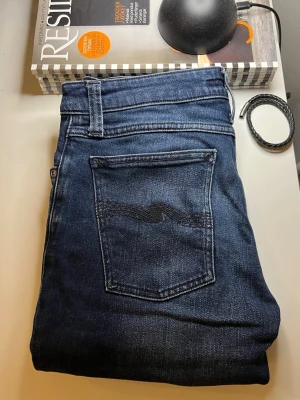 Mörkblå jeans från Nudie Jeans - Snygga mörkblå jeans från Nudie Jeans med klassisk femficksdesign och smal passform. Jeansen har den ikoniska vågade sömmen på bakfickorna och är tillverkade i denim med lätt stretch för extra komfort.
