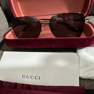 Svarta solglasögon från Gucci - Snygga svarta solglasögon från Gucci med rektangulära linser och tunna metallbågar. Skalmarna har en röd och grön detalj som ger en exklusiv känsla. Kommer i ett lyxigt vinrött fodral med Gucci-logga.