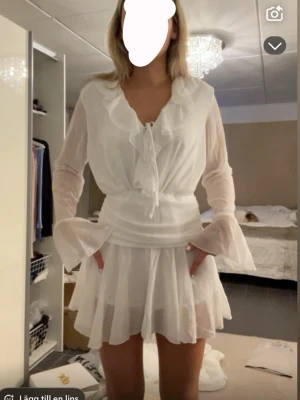 Student klänning  - Söt vit kortklänning med volangdetaljer och omlottknytning framtill. Klänningen har långa, lätt transparenta ärmar med utsvängda ärmslut och en flowy kjol. Perfekt för sommarens alla tillfällen.