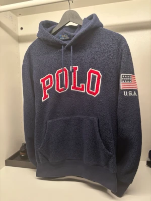 Marinblå fleece hoodie Polo Ralphlauren  - Mysig marinblå hoodie från Polo Ralph Lauren i fleece med stor röd och vit POLO-logga på bröstet och amerikansk flagga med U.S.A. på ärmen. Klassisk känguruficka och huva med snörning. Perfekt för dig som gillar streetwear vibes. Väldigt snyggt, unikt och sällsynt plagg! Otroligt bra skick. Har du några frågor så fråga gärna, jag svarar snabbt!🙌🔥
