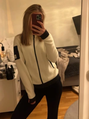 Vit fleecejacka från Peak Performance - Säljer en vit fleecejacka från Peak Performance med svarta detaljer. Jackan har hel dragkedja, svarta muddar och fickor med dragkedja. Peak Performance-logga på ärmen. Perfekt för lager-på-lager och chill vardagsstil.