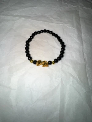 Svart pärlarmband med gulddetalj - Snyggt armband med svarta pärlor och guldfärgade detaljer, inklusive en cool guldfärgad drakfigur i mitten. Perfekt accessoar för att lyfta din stil och ge en unik touch till din outfit. Elastiskt band gör att det passar de flesta handleder.