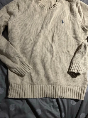 Beige stickad tröja Polo Ralph Lauren - Klassisk beige stickad tröja från Polo Ralph Lauren med rund halsringning och ribbade muddar. Tröjan har en liten blå broderad logga på bröstet och är tillverkad i mjukt material. Perfekt för lager-på-lager och stilren look. Använd bara ett fåtal par gånger