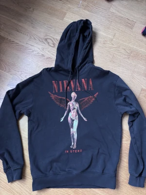 Svart Nirvana In Utero hoodie - Svart hoodie med tryck av Nirvanas ikoniska In Utero-albumomslag på bröstet. Stor röd text med bandnamnet och motiv av en änglalik figur. Klassisk huva med dragsko och ribbade muddar. Perfekt för dig som älskar grunge och vill ha en statement-piece. Använd sparsam. Den är grå. Inga fel. 