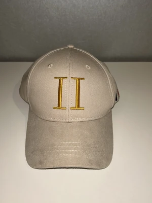 Beige keps med gul brodyr II - Snygg beige keps med gul broderad II-logga framtill. Klassisk böjd skärm och justerbar rem bak. Tillverkad i bomullstyg som ger en avslappnad och stilren look. Perfekt accessoar för att lyfta din outfit.
