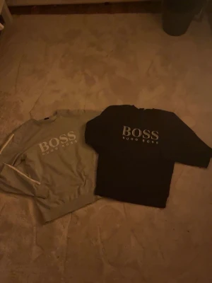 2 BOSS Hugo Boss sweatshirts - Två stilrena sweatshirts från Hugo Boss, en i grått och en i svart. Båda har klassisk rund hals och stor BOSS-logga framtill. Perfekta för en avslappnad och trendig look. Mjukt material och lång ärm.