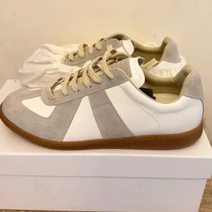 Maison Margiela Replica sneakers - Stilrena Maison Margiela Replica sneakers i vitt och ljusgrått läder och mocka. Klassisk låg modell med beige snörning och brun gummisula. Perfekta för dig som gillar minimalistisk och tidlös design.
