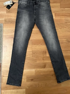 Grå slim jeans från Jack and Jones - Snygga grå jeans från Jack and jones i slim fit-modell. Jeansen har klassisk femficksdesign, svarta knappar och en tvättad look med ljusare partier framtill. Perfekta för en trendig och avslappnad stil.
