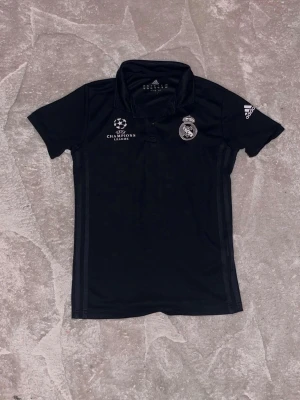 Svart Real Madrid pikétröja Adidas - Svart pikétröja från Adidas med Real Madrid-logga och Champions League-tryck på bröstet. Tröjan har korta ärmar, krage och knappar framtill. Perfekt för fotbollsfans som vill ha en sportig och stilren look.