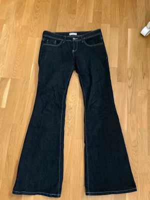Mörkblå bootcut jeans med vita sömmar - Snygga mörkblå bootcut jeans med kontrasterande vita sömmar och klassiska fem fickor. Jeansen har låg midja och utsvängda ben, perfekt för en trendig och avslappnad look. Materialet är denim som ger en cool och tidlös känsla. Köpta på Gina, storlek 38
