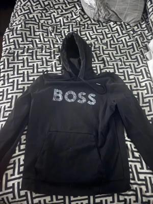 Svart hoodie från BOSS med tryck - Svart hoodie från BOSS med stort mönstrat BOSS-tryck på bröstet. Klassisk modell med huva och dragsko samt en stor magficka framtill. Perfekt för en avslappnad och cool stil. Använd fåtal gånger.
