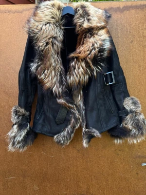 Svart skinnjacka med äkta päls - Säljer en svart skinnjacka med stora, fluffig päls i bruna och beige toner på krage och ärmslut. Jackan har bälte med spänne i midjan och vid handlederna för extra edge. Perfekt för dig som vill sticka ut och hålla dig varm med stil.