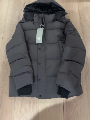 Mörkgrå dunjacka från Canada Goose - Snygg mörkgrå dunjacka från Canada Goose med svart logotyp-patch på ärmen. Jackan har huva, raka kanaler och svarta muddar vid ärmsluten. Perfekt för kalla vinterdagar och riktigt varm tack vare dunfyllningen.