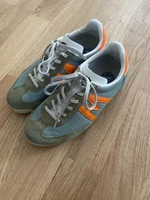 Karhu sneakers i grönt och orange - Snygga sneakers från Karhu med gröna mockadetaljer, mesh i ljusgrönt och orangea inslag på sidor och häl. Klassisk rund tå och snörning framtill. Perfekta för dig som gillar retrostil och vill sticka ut med färg.