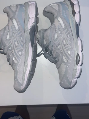 Gråa sneakers från Asics - Snygga gråa Asics sneakers med mesh och syntetdetaljer. Skorna har en sportig vibe med klassisk snörning och en robust sula i grått och ljusrosa. Perfekta för dig som gillar streetstyle och bekväma skor.