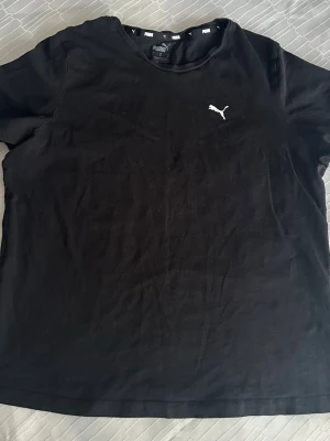 Svart t-shirt från Puma - Svart t-shirt från Puma med klassisk rund hals och vit Puma-logga på bröstet. Tillverkad i mjuk bomull som känns skön mot huden. Perfekt för en avslappnad och sportig stil.