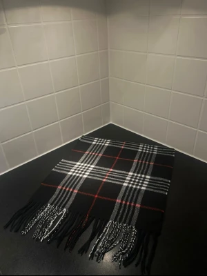 Svart rutig halsduk med fransar - Stilren svart halsduk med klassiskt rutmönster i vitt och rött. Halsduken har fransar i ändarna och är perfekt för att ge din outfit en snygg touch. Passar både till jacka och kappa. Materialet känns mjukt och bekvämt mot huden. Likade burberry 