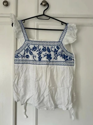 Vit blus med blå broderi och volang - Supersöt vit blus med blått broderat blommönster upptill och volangdetaljer på axlarna. Blusen har fyrkantig ringning, breda axelband och knytband i sidorna. Perfekt för sommarens alla tillfällen!