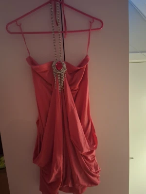 Rosa kortklänning med smyckesdetalj - Säljer en rosa kortklänning med tunna axelband och draperad kjol. Klänningen har en stor dekorativ smyckesdetalj framtill med rosa sten och hängande strass. Perfekt för dig som vill sticka ut med en unik look. Säljes billigt för några stenar har lossnat men inget som syns